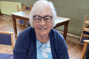 Photo of a senior woman with glasses smiling at the camera. Shoulders upwards.