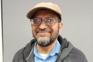 Headshot of a man with a hat and glasses smiling at the camera. Shoulders upwards.