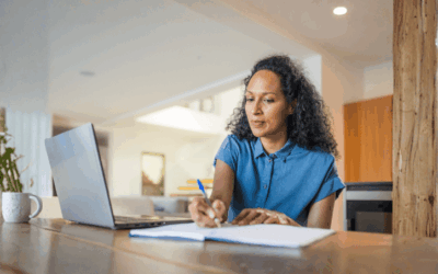 Woman sat at her dining room table with a laptop in front of her. She's taking notes